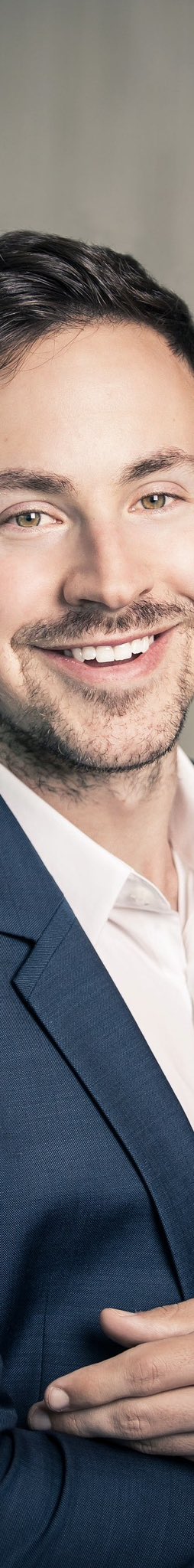 A close-up portrait of a smiling young man with dark hair, light stubble, and hazel eyes, wearing a navy blue blazer over a white collared shirt.