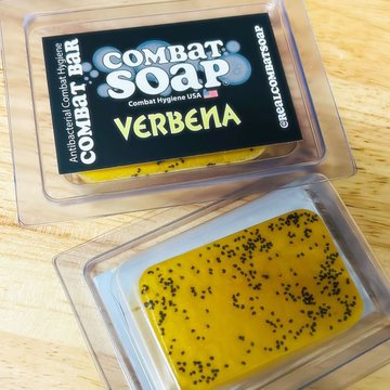 Two bars of Combat Soap® in "Verbena" scent are displayed in clear plastic packaging, with one bar shown face-down revealing its yellow surface embedded with black seeds. The image introduces the first product arriving in the MONOLITH Merch Store—a combat athlete hygiene brand founded in 2011 that the post announces is now coming onto the XRPL.