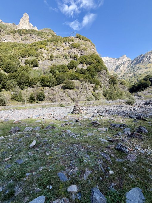 A scenic mountain landscape featuring a rocky, dry riverbed in the foreground, leading up to steep, forested mountains under a blue sky.