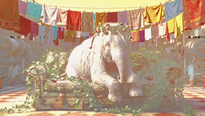 A white elephant rests on a wooden platform surrounded by greenery, with colorful fabrics and tapestries hanging on lines in the background.
