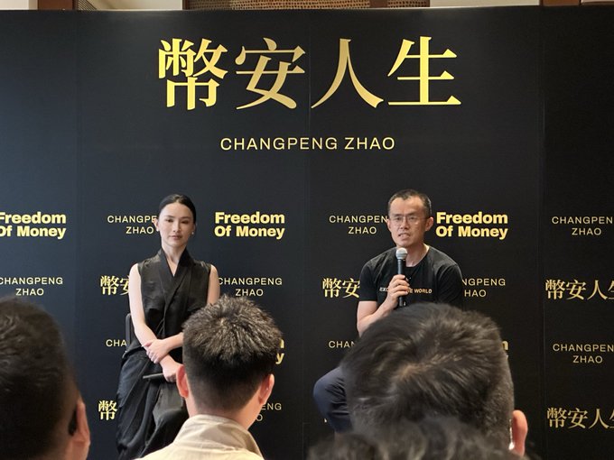 Changpeng Zhao (CZ) speaks into a microphone while seated on a stage at an event branded 'Freedom Of Money' with Chinese characters '幣安人生' behind him.