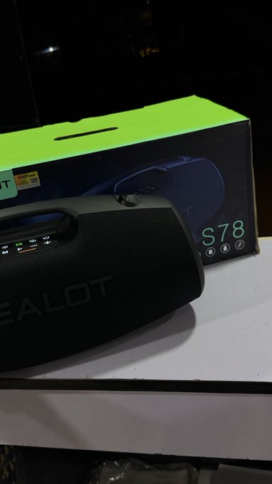 A black portable Bluetooth speaker branded 'ZEALOT' sitting on a white box, with its green and black packaging visible behind it.
