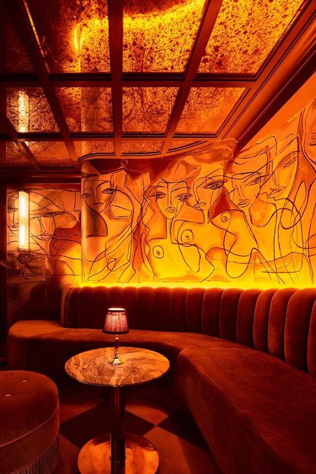 A dimly lit, atmospheric lounge or bar interior with warm orange/red lighting, featuring abstract line-art murals of faces on the walls, a curved velvet banquette seating, and a small round marble table with a lamp.