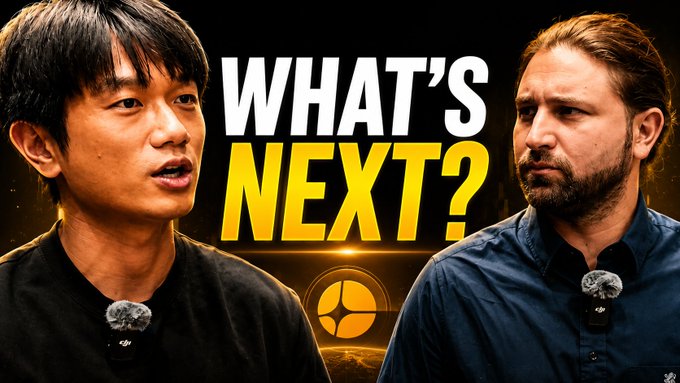 The image features two men standing next to each other, with one man wearing a black shirt and the other wearing a blue shirt. They are both talking into microphones, suggesting that they might be discussing something related to cryptocurrency or blockchain technology. The background of the image is dark gray, which contrasts with the white text on the image. This contrast makes it easy for viewers to read and understand the content displayed in the image.