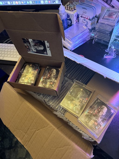 An overhead view of an open cardboard box on a desk filled with stacks of foil-packaged trading cards, some featuring bear imagery, alongside computer monitors and a keyboard.