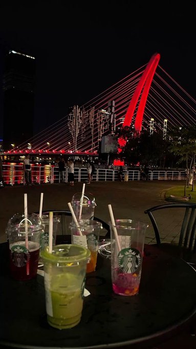 Starbucks iced beverages on a table overlooking a cityscape and a red-illuminated bridge at night.