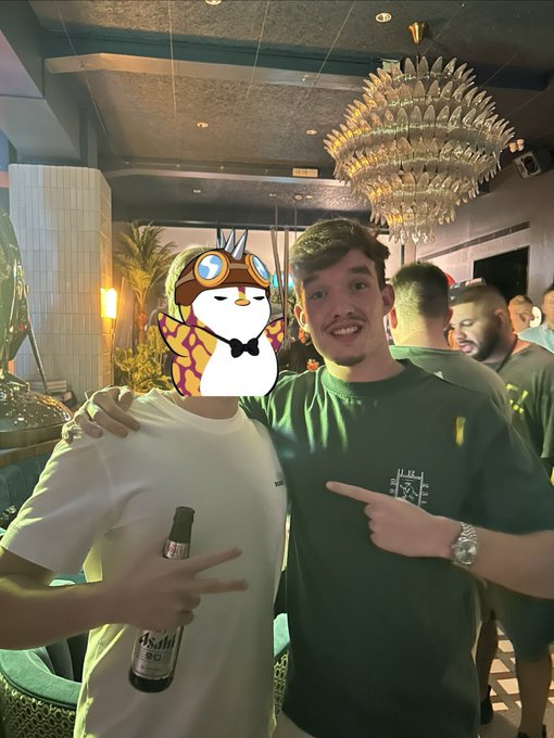 Two men pose together at an indoor event with a chandelier, one holding a bottle of Asahi beer, while the face of the man on the left is covered by a penguin sticker.