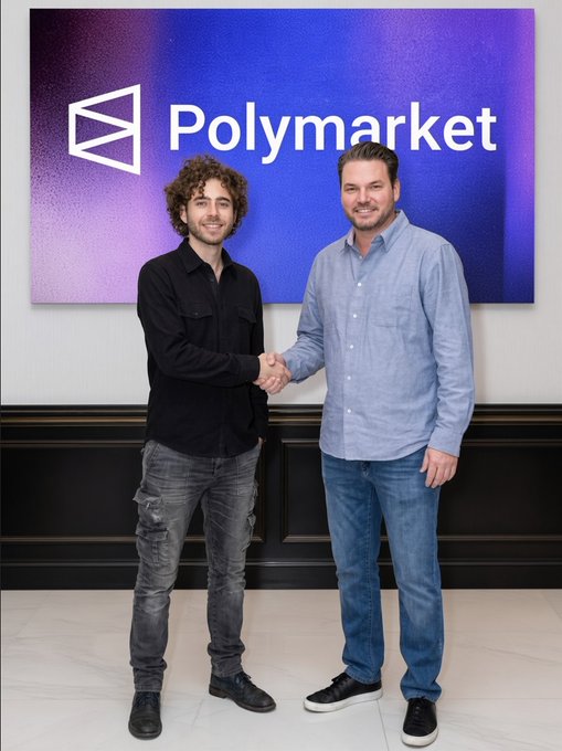 Two men shaking hands in front of a large 'Polymarket' logo display, appearing to be in a corporate or partnership setting with marble flooring.