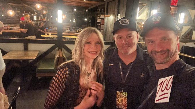 Three people smile for a photo inside a restaurant/bar, with two men wearing black baseball caps and a blonde woman in a black mesh top.