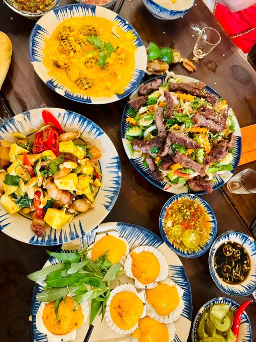 An overhead view of a wooden dining table spread with various Vietnamese dishes, including a yellow curry with stuffed items, stir-fried beef with vegetables, a tofu and vegetable dish, fried snacks, and small bowls of dipping sauces.