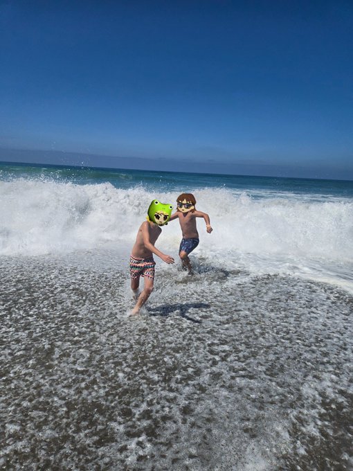 Two children in swim trunks wearing animal-themed helmets (one green frog, one monkey) running playfully through ocean waves at a beach under a clear blue sky.