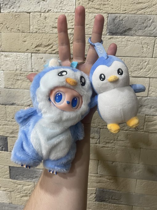A hand holding two blue and white penguin-themed plush keychains, one larger wearing a hooded costume and a smaller simpler one.