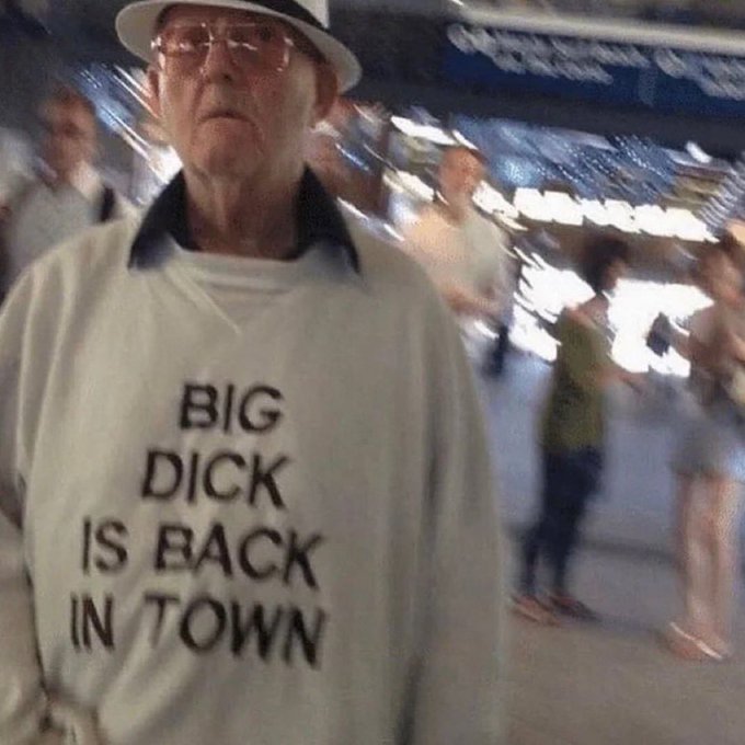 An older man wearing a white hat and glasses stands in a crowd, wearing a white sweatshirt with bold text reading 'BIG DICK IS BACK IN TOWN'.
