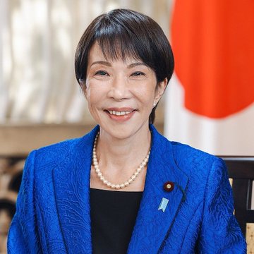 An official portrait of a Japanese woman with short black hair, wearing a blue textured blazer over a black top, accessorized with a pearl necklace and a blue ribbon pin, posed in front of the Japanese flag.
