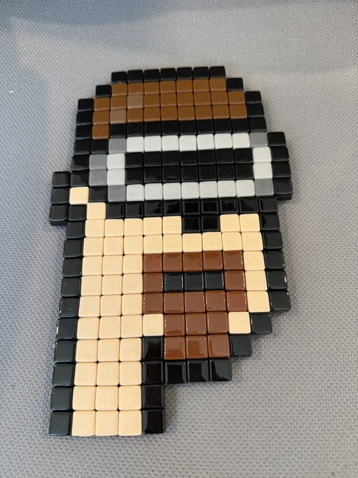 A pixel art character made from small square tiles in black, brown, white, and tan colors, depicting what appears to be a figure with a hat and face details.