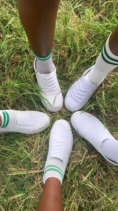 Four pairs of feet stand in a circle on green grass, all wearing matching white sneakers and white tube socks with green stripes.