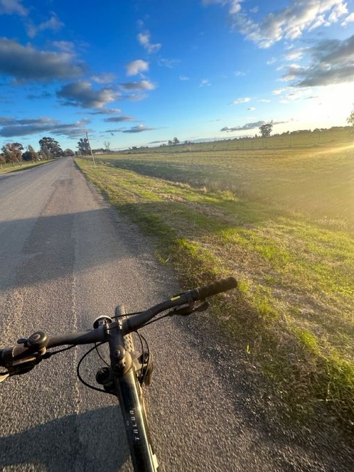 A first-person view of cycling down a rural road at sunset, with the bicycle's handlebars visible in the foreground against a grassy landscape.