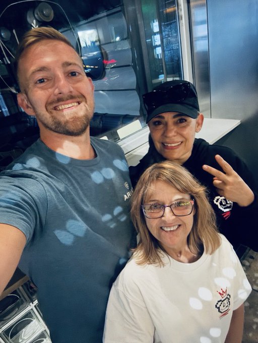 A selfie of three smiling people—a man with a beard, a woman in a black cap making a peace sign, and a woman with glasses and blonde hair—taken indoors near a large window.