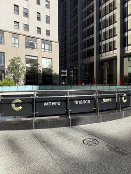 An outdoor urban scene shows a building entrance with a glass railing displaying the text "where finance flows" alongside a stylized "C" logo, located in front of a multi-story building.