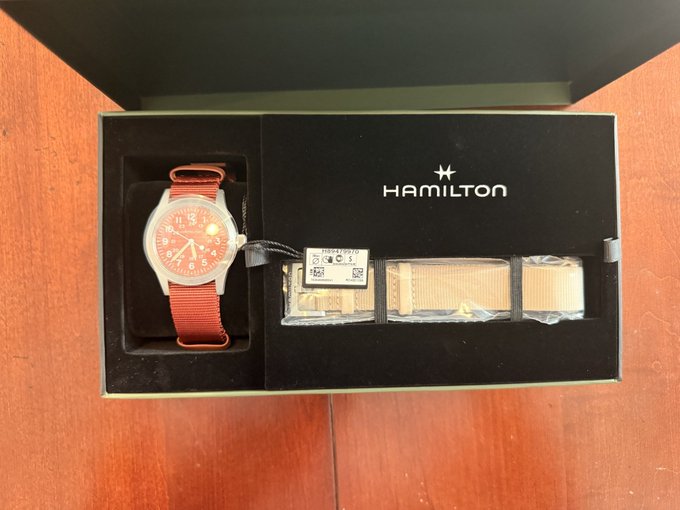 A Hamilton watch with a red dial and matching red strap sits in its presentation box alongside additional beige straps, including one labeled "H70455580," as a gift or prize related to the Magic Internet Math podcast discussion on Bitcoin's libsecp256k1 cryptography.