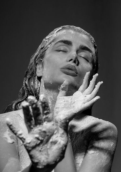 A black and white artistic portrait shows a woman with closed eyes, her face and hands covered in what appears to be a white textured substance like clay or paint.