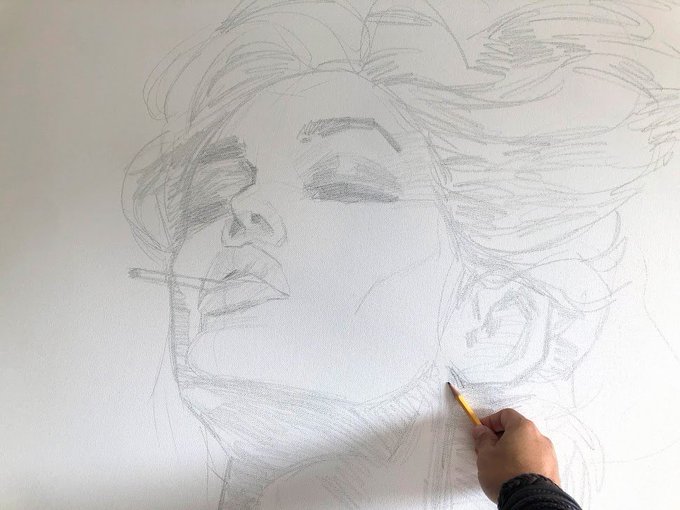 A hand holds a pencil, sketching the detailed face and flowing hair of a portrait on white paper.