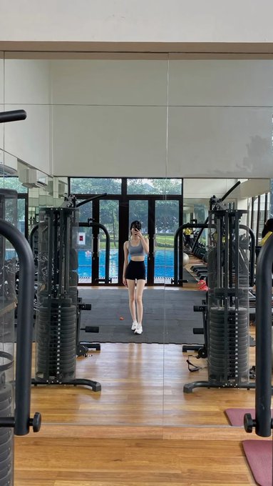 A woman in workout clothes takes a mirror selfie in a gym with weight machines and a pool visible in the background.