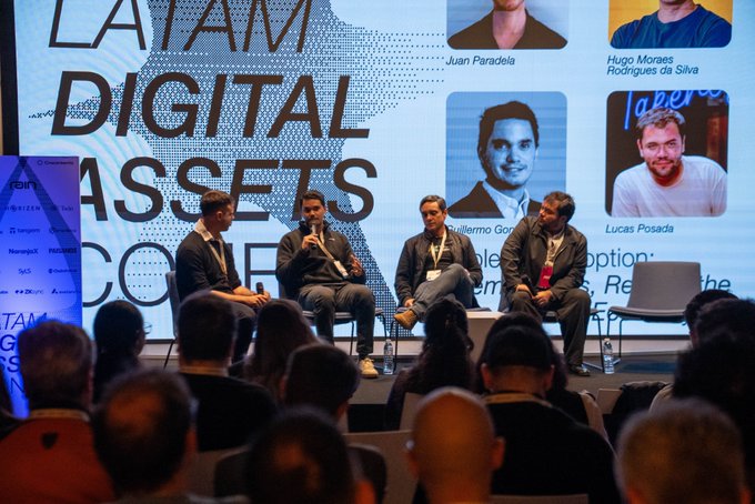 Four men sit on a panel discussion at the 'LATAM Digital Assets Conference,' with a large screen behind them showing headshots of speakers including Juan Paradela, Hugo Moraes Rodrigues da Silva, Guilherme Gor (Guilherme Gores), and Lucas Posada.