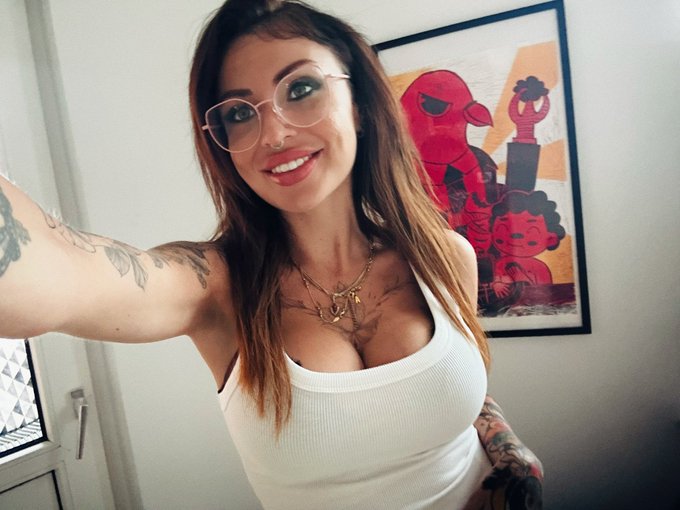 A woman with long brown hair, glasses, and neck tattoos takes a selfie while wearing a white tank top.