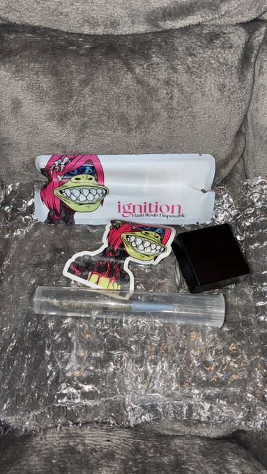 An 'ignition' brand stash kit laid out on bubble wrap and gray fabric, containing a rolling tray, grinder, glass tube, stickers featuring Pepe the Frog designs, and a black container.