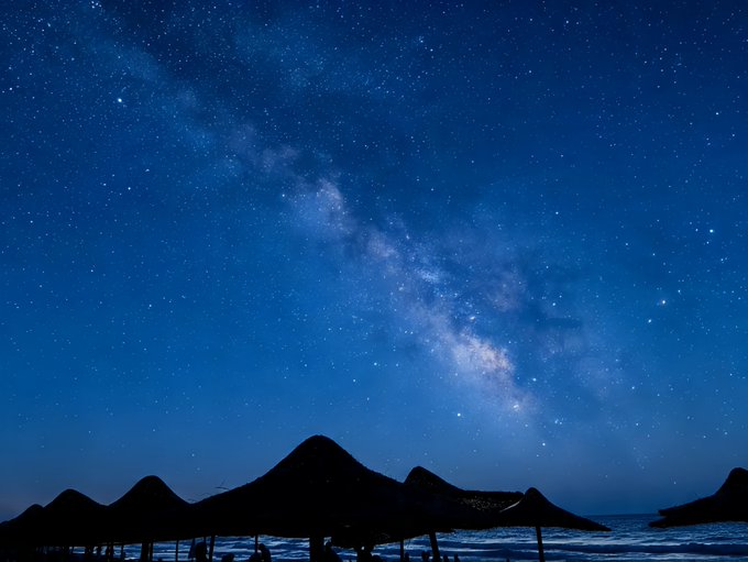 A nighttime photograph showing the Milky Way galaxy stretching across a starry blue sky above the dark silhouette of a mountain and beach with umbrellas.
