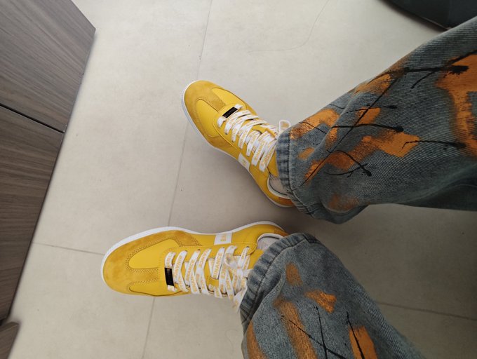 An overhead view of someone wearing bright yellow sneakers with white and black striped laces, paired with blue jeans featuring an orange pattern, standing on a tiled floor.