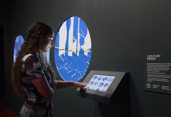 The image shows a woman with curly hair standing next to a display of modern art, which includes a large screen or monitor that displays various images. The woman appears to be looking at one of these screens, possibly reading about the artwork displayed on it. The setting seems to be an indoor space dedicated to showcasing contemporary pieces and providing information about them.