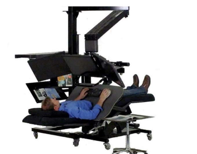 A man in a blue shirt lies fully reclined in an elaborate, multi-monitor workstation, working from a near-horizontal position while typing on a keyboard balanced on his chest. The "trenches" reference humorously comes through—the image exaggerates the sedentary, immovable reality of deep computer work, where the user has essentially melted into the desk itself rather than ever leaving it.