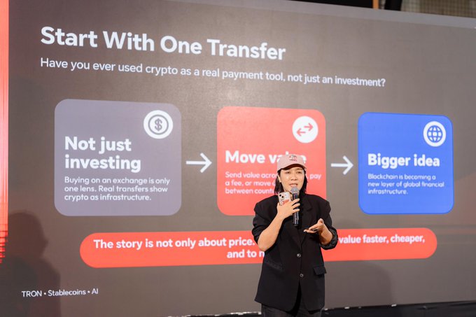 A presenter in a black blazer stands on stage holding a microphone in front of a large presentation screen titled 'Start With One Transfer' discussing crypto as payment, with TRON and Stablecoins branding visible.