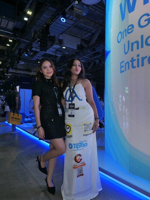 Two women stand together at what appears to be a crypto/tech conference, with one wearing a long white dress covered in sponsor logos including "Trilorp," "bagel," "WEEX," and a QR code. A large illuminated screen to the right displays partial text reading "W... one... Unlo... Entire..." against a bright blue background, suggesting a branded event backdrop.