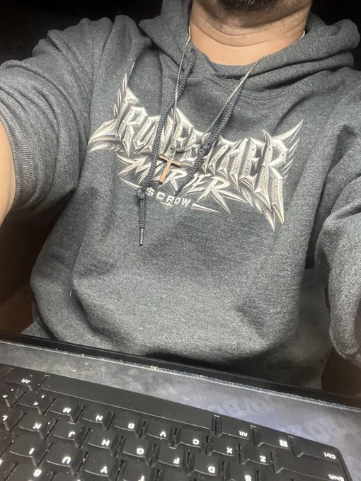 A person is shown from the chest down, wearing a gray hoodie with a metallic 'Crown The Empire' graphic, a cross necklace, and sitting behind a black keyboard.