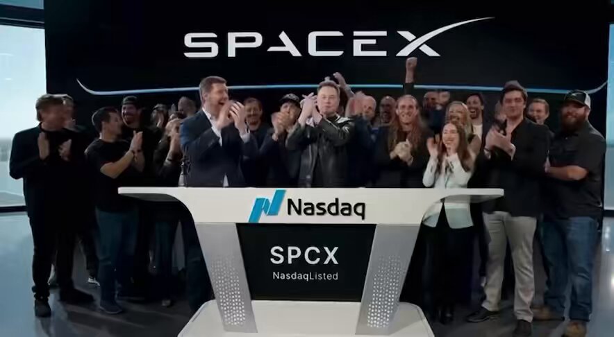 The image features a group of people standing around a large desk, with some of them clapping their hands together. The scene appears to be set at a company event or gathering, possibly related to the announcement of a new company or the launch of a new product. In the background, there is a sign that reads "SPACEX" and "Nasdaq", indicating that this event is taking place in front of a company's headquarters. The group consists of people standing on both sides of the desk, with some individuals closer to the foreground and others further back.