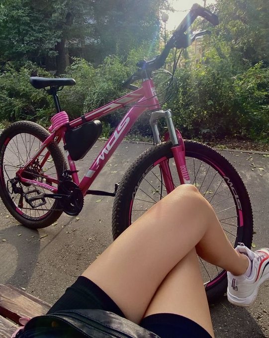 A pink EMC mountain bike leans against a bench in a park setting, with a person's legs visible in the foreground wearing shorts and white FILA sneakers.