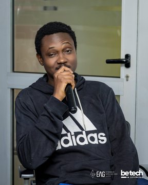 A man wearing a black Adidas hoodie holds a microphone while seated, with logos for 'EAC' and 'be tech' visible in the bottom right corner.
