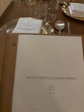 A close-up of a menu for an "ARCUS X BOOGLE X BAXUS DINNER" sits on a set dining table with glassware, aligning with the post about a collaborative NYC event hosted by Arcus, Boogle, and BAXUS.