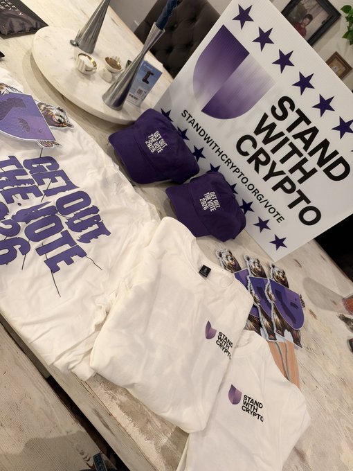 A display table showing merchandise for the 'Stand With Crypto' campaign, including t-shirts, hats, and stickers featuring the campaign logo and the URL 'STANDWITHCRYPTO.ORG/VOTE'.