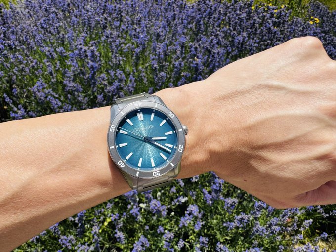 A wrist shot displays a blue-dial wristwatch (appears to be an Omega with "OMEGA" text visible on the face) set against a backdrop of lavender flowers. The luxury timepiece complements the post's theme of "collecting Azuki grails" by showcasing another prized acquisition, much like the sought-after elements referenced in the popular phrase.

**Visible text/numbers:** "OMEGA" on the watch dial, and numerals 10, 20, 30, 40, 50 on the bezel.