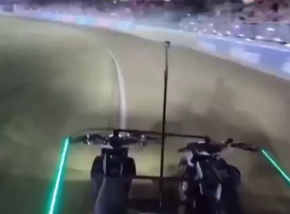 The image shows a motorcycle race taking place at night, with two motorcycles racing around a track. One of the motorcycles has green lights on its wheels, which are likely used for visibility purposes during nighttime races. The other motorcycle also has green lights on its wheels. In the background, there is a crowd watching the race from the stands, and the image includes text that reads "Ðoge is battling with ." and "Ðoge is battling with .08¢ again today."