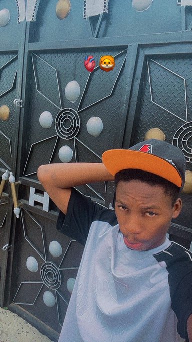 A young man wearing a white and black t-shirt and a black and orange baseball cap poses with his arm raised in front of a dark metal gate decorated with circular patterns.