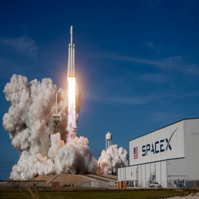 A SpaceX Falcon Heavy rocket launching from the pad with massive smoke clouds, with the 'SPACEX' logo visible on the building to the right.
