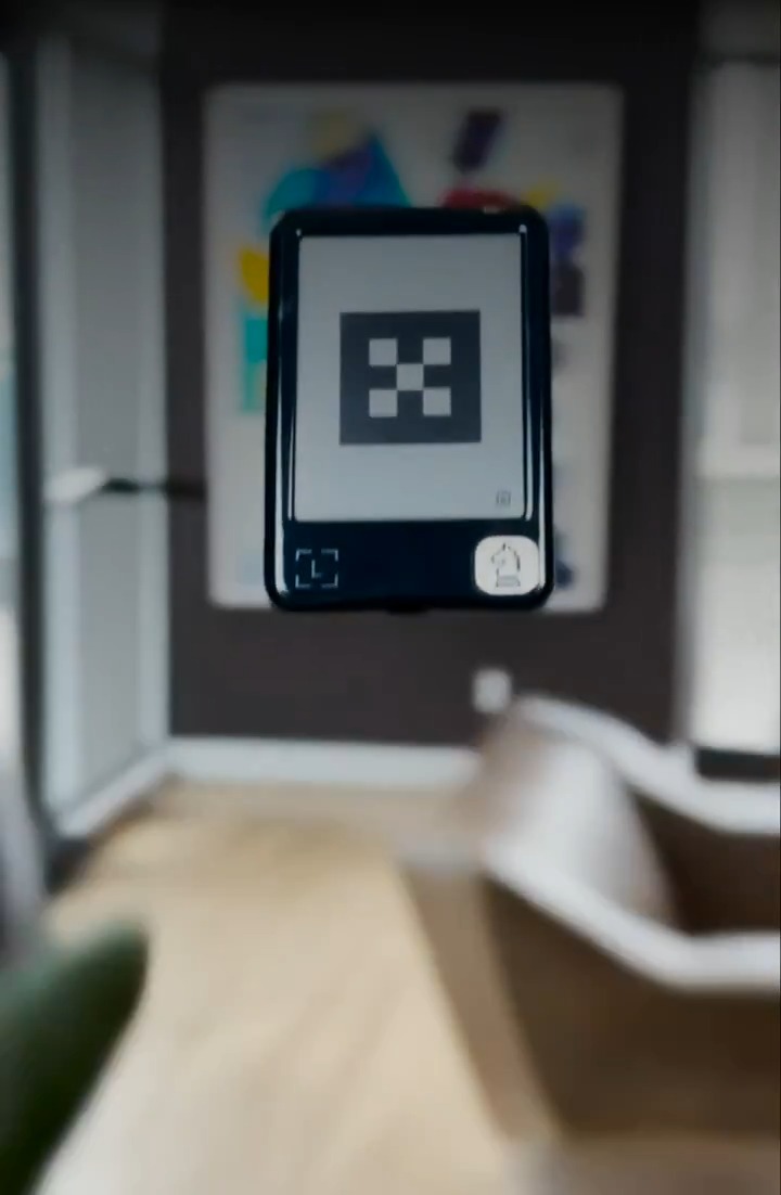 A handheld device displaying a QR code and a bell icon is held up in focus against a blurred background of a room with a door and abstract artwork.