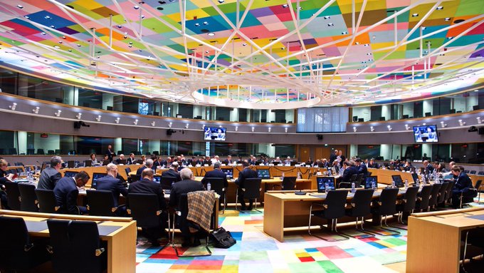 Interior view of a large meeting room with curved tiered seating filled with people, featuring a colorful geometric patterned ceiling and floor in the European Parliament style.