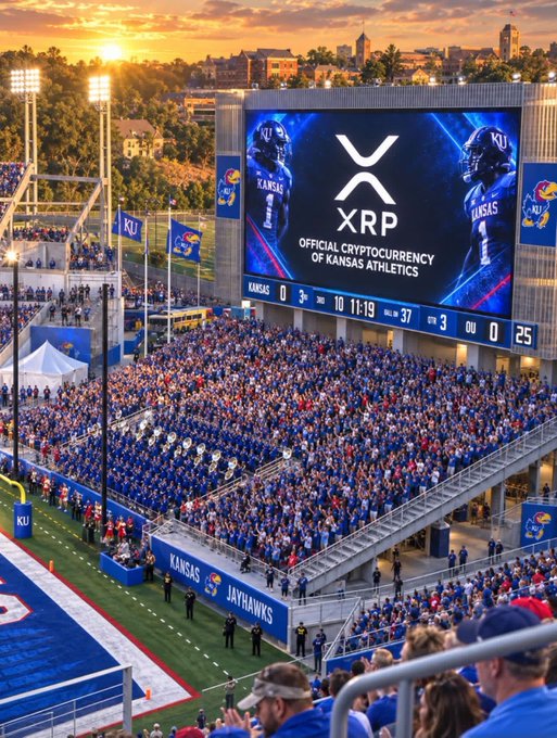 A Kansas Jayhawks football stadium filled with fans wearing blue, with a large scoreboard displaying XRP as the 'Official Cryptocurrency of Kansas Athletics' alongside images of players, during a sunset game.