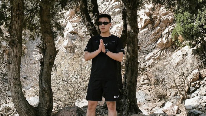 A young man stands outdoors among trees and rocky terrain, wearing sunglasses and a black OKX-branded t-shirt and shorts, with hands pressed together in a prayer-like gesture. The image complements the post's playful theme of "praying" for Dogecoin, showcasing the OKX merchandise as the "battle robe" mentioned in the caption.